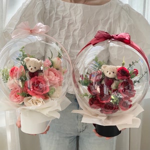 May include: Two clear glass globes filled with pink and red roses and teddy bears. The globes are sitting on white and black bases.