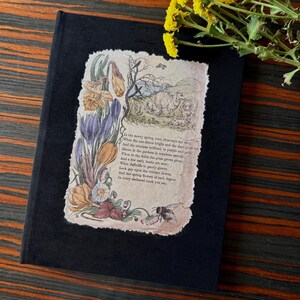 May include: A black journal with a vintage-style illustration of spring flowers and a pastoral scene. The illustration features daffodils, crocus, and a bee, with text from a poem. The journal is a hardcover book.