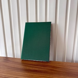 Puede incluir: Un libro de tapa dura con una cubierta verde oscuro descansa sobre una superficie de madera. La cubierta del libro es de un color verde oscuro s&oacute;lido, y las p&aacute;ginas son visibles a lo largo del borde. El fondo presenta una pared blanca con listones verticales.