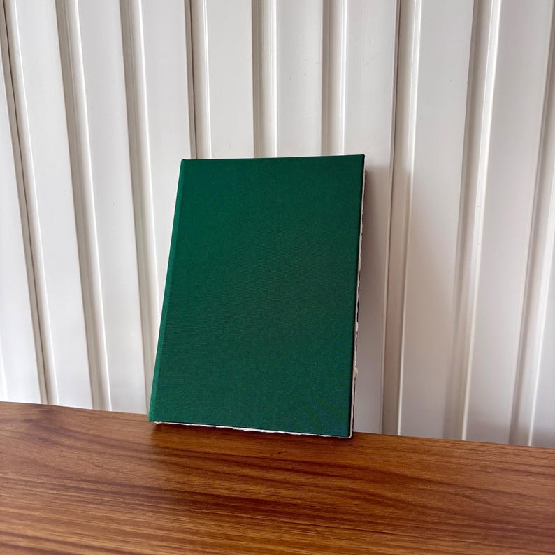 Puede incluir: Un libro de tapa dura con una cubierta verde oscuro descansa sobre una superficie de madera. La cubierta del libro es de un color verde oscuro s&oacute;lido, y las p&aacute;ginas son visibles a lo largo del borde. El fondo presenta una pared blanca con listones verticales.