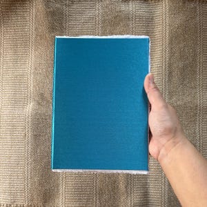 May include: A teal blue notebook with a white border. The notebook is lying flat on a brown textured surface.