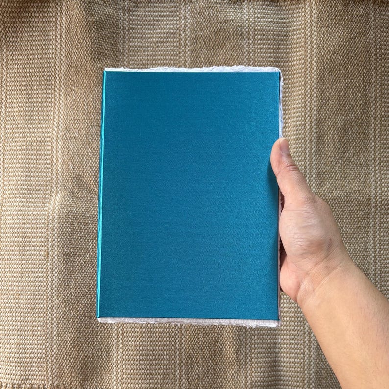 May include: A teal blue notebook with a white border. The notebook is lying flat on a brown textured surface.