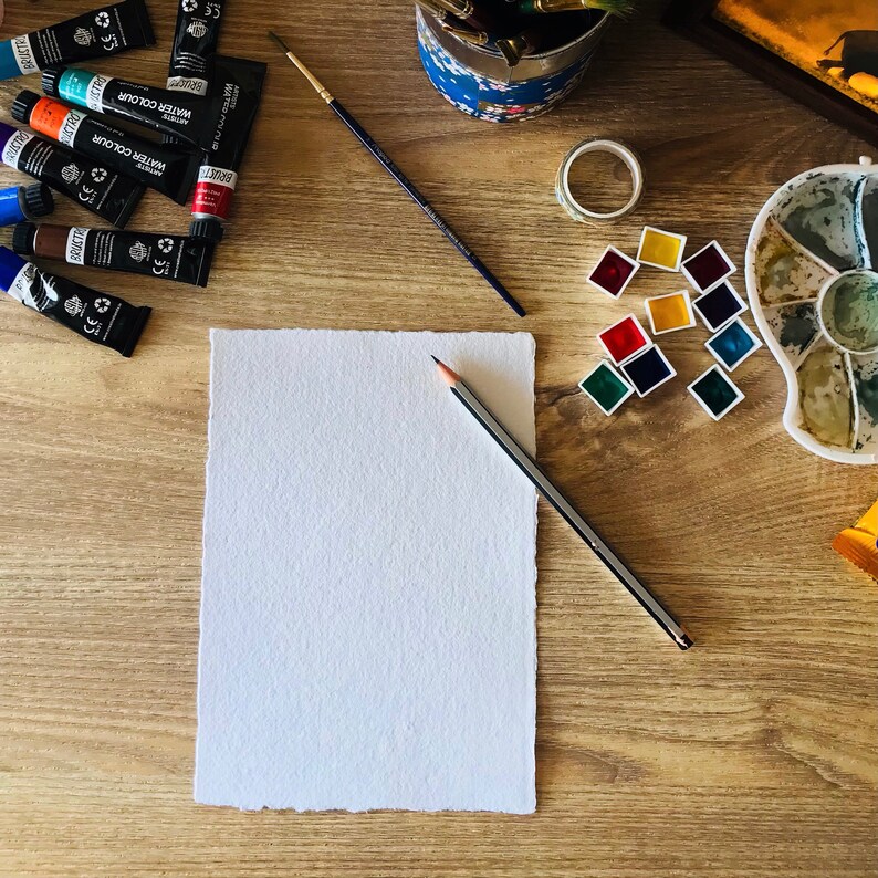 May include: A white sheet of paper with a pencil on a wooden table. There are watercolor paints, a paintbrush, and a roll of tape on the table.