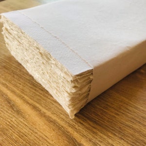 K&ouml;nnte beinhalten: Ein Stapel wei&szlig;er handgemachter Papierb&ouml;gen, gebunden mit einem wei&szlig;en Papierumschlag. Das Papier hat eine raue, strukturierte Oberfl&auml;che.