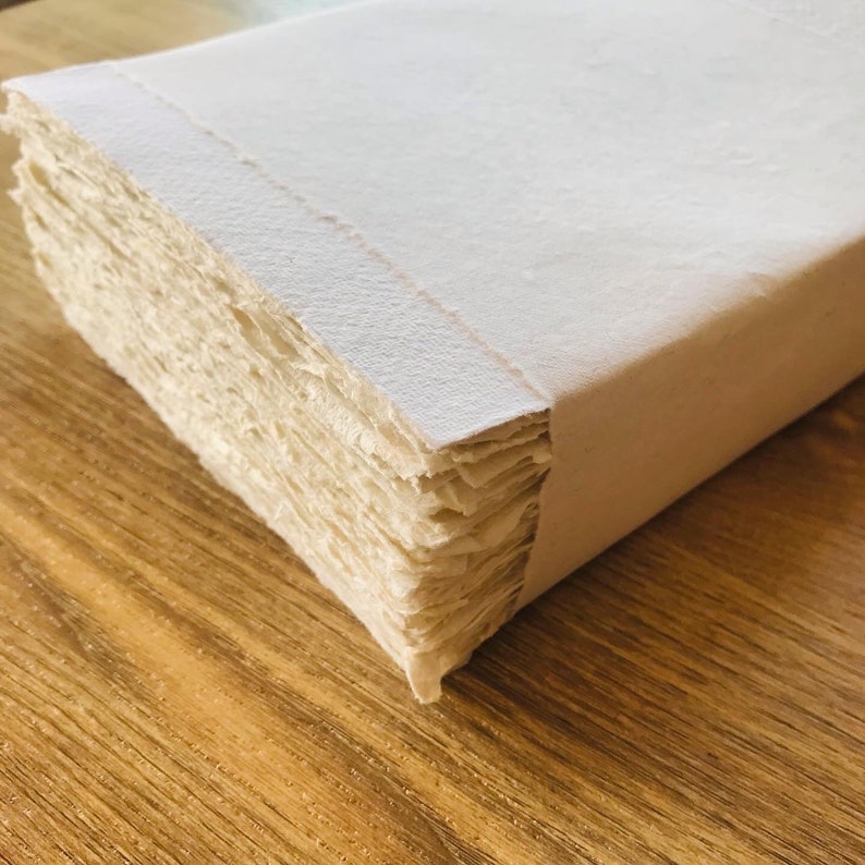 K&ouml;nnte beinhalten: Ein Stapel wei&szlig;er handgemachter Papierb&ouml;gen, gebunden mit einem wei&szlig;en Papierumschlag. Das Papier hat eine raue, strukturierte Oberfl&auml;che.