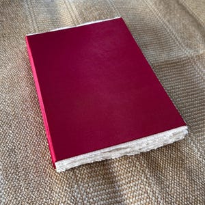 May include: A red hardcover journal with white, textured paper edges. The journal is closed and lying on a brown, textured surface.