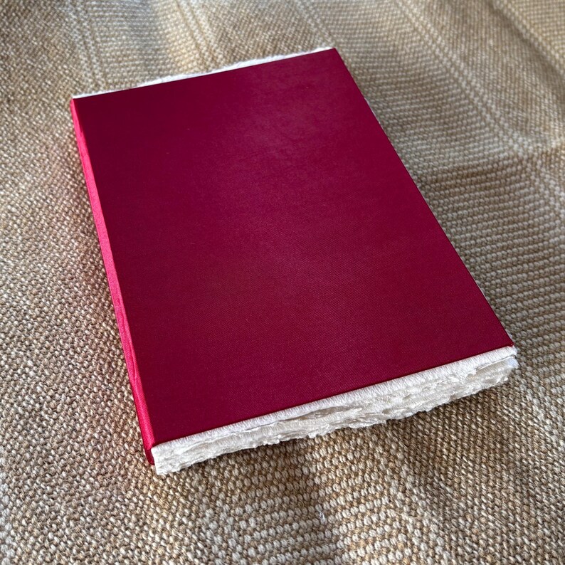 May include: A red hardcover journal with white, textured paper edges. The journal is closed and lying on a brown, textured surface.