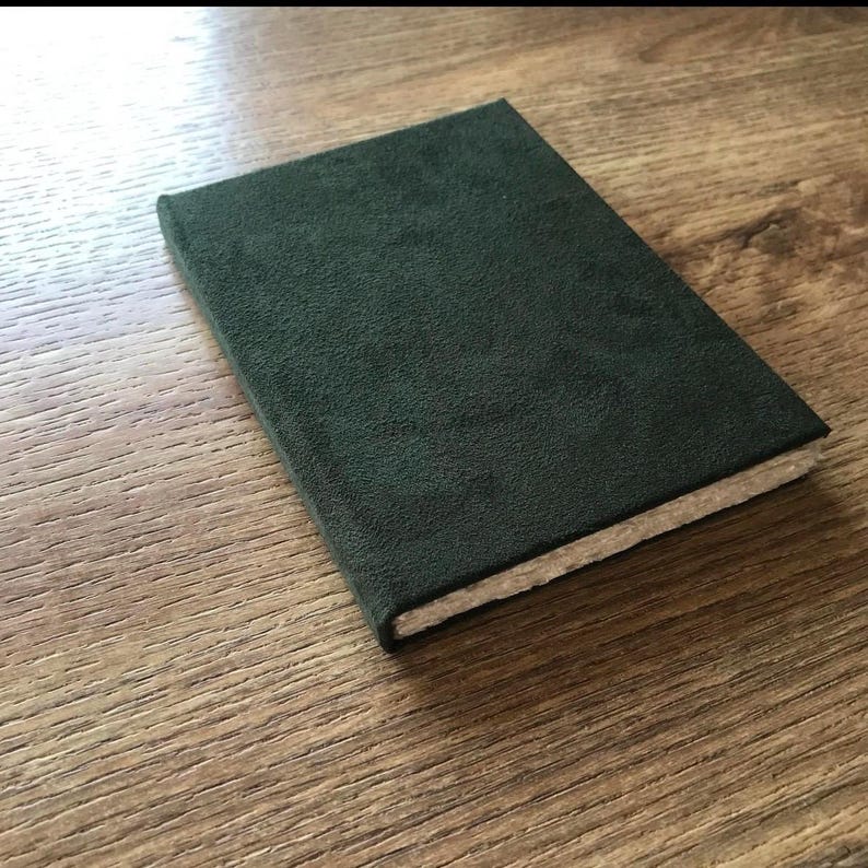 Op de afbeelding: Een donkergroen notitieboek met een getextureerde kaft. Het rechthoekige notitieboek heeft een zacht, su&egrave;de-achtig uiterlijk. De pagina's zijn zichtbaar langs de rand en het ligt op een houten oppervlak.