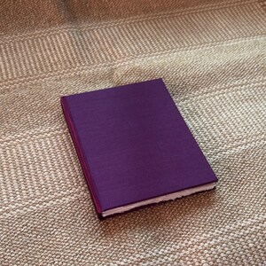 Op de afbeelding: Een gesloten, rechthoekig notitieboek met een dieppaarse kaft ligt op een getextureerd, beige oppervlak. De pagina's van het notitieboek zijn zichtbaar langs de open rand en de rug is een donkerdere tint paars.