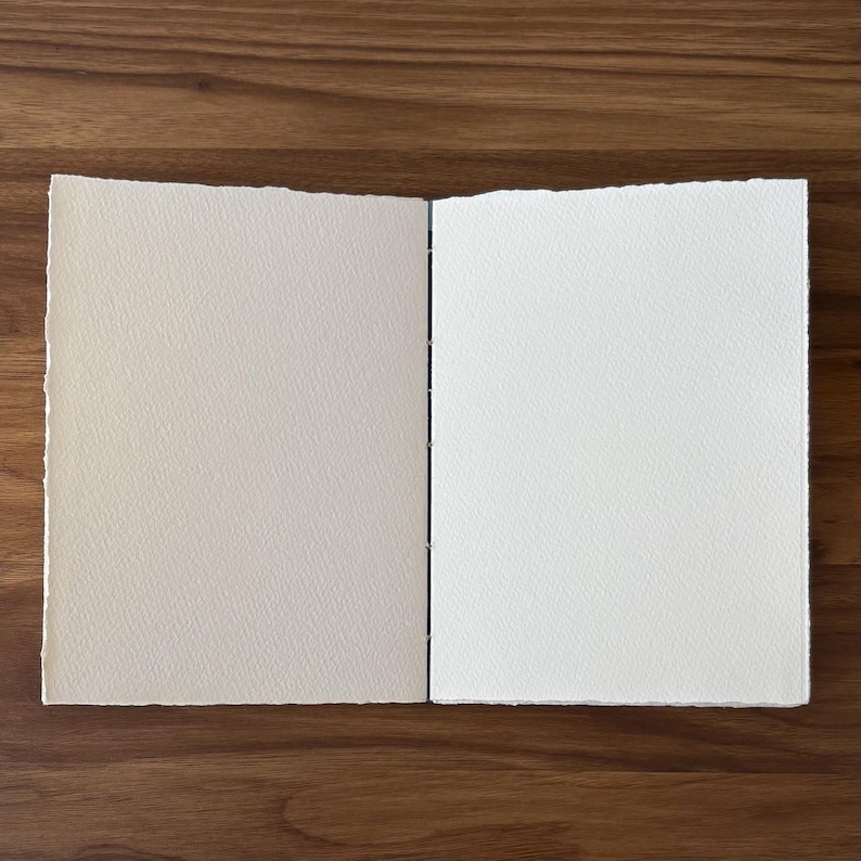 K&ouml;nnte beinhalten: Ein offenes, handgefertigtes Journal mit strukturierten, cremefarbenen Seiten. Das Journal ist mit sichtbaren N&auml;hten am R&uuml;cken gebunden. Die Seiten haben einen B&uuml;ttenrand, und das Journal liegt auf einer dunklen Holzoberfl&auml;che.