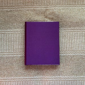 Op de afbeelding: Een rechthoekig boek met een dieppaarse kaft. Het boek ligt gesloten op een beige, getextureerd oppervlak. De randen van het boek zijn een donkerdere tint paars.