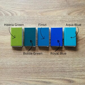 May include: Five book cover shades arranged in a row on a wood surface. The shades are labeled 'Heena Green', 'Bottle Green', 'Firozi', 'Royal Blue', and 'Aqua Blue'.
