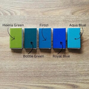 May include: Five small, rectangular colour swatches in shades of green and blue are arranged on a wooden surface. The colours are labelled: Heena Green, Bottle Green, Firozi, Royal Blue, and Aqua Blue. Each swatch has a white border.