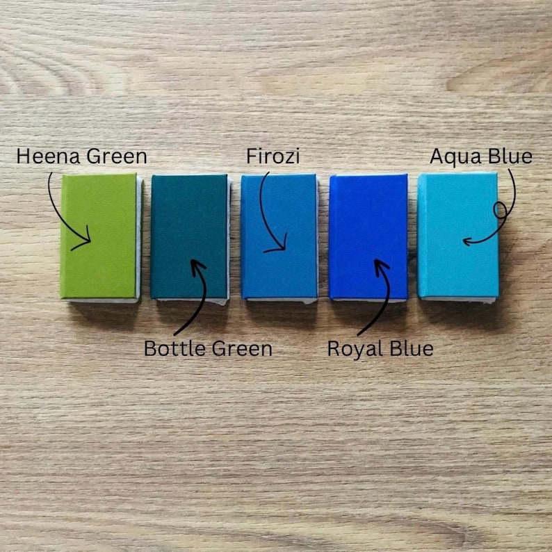 May include: Five small, rectangular colour swatches in shades of green and blue are arranged on a wooden surface. The colours are labelled: Heena Green, Bottle Green, Firozi, Royal Blue, and Aqua Blue. Each swatch has a white border.