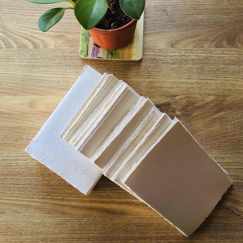 May include: A stack of white handmade paper sheets with deckled edges. The paper is arranged in a fan shape, with the top sheet folded over the rest. The paper is on a wooden surface.