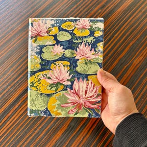 K&ouml;nnte beinhalten: Ein Notizbuch mit floralem Seerosenmuster. Der Einband zeigt rosa Seerosen, gelbe Seerosenbl&auml;tter und gr&uuml;ne Bl&auml;tter auf einem dunkelblauen Hintergrund. Das Notizbuch wird in einer Hand gehalten.