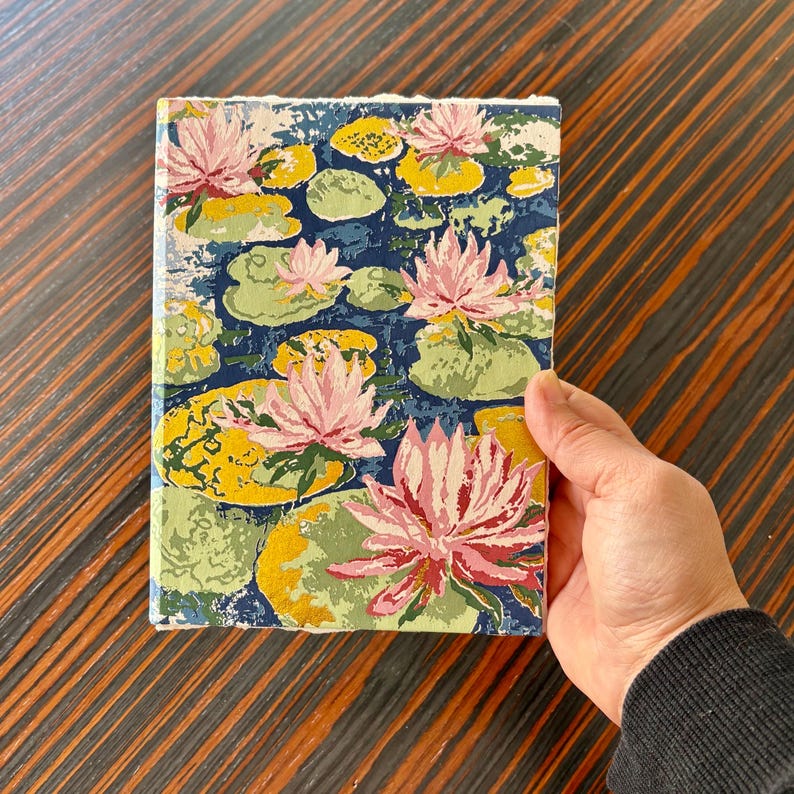 K&ouml;nnte beinhalten: Ein Notizbuch mit floralem Seerosenmuster. Der Einband zeigt rosa Seerosen, gelbe Seerosenbl&auml;tter und gr&uuml;ne Bl&auml;tter auf einem dunkelblauen Hintergrund. Das Notizbuch wird in einer Hand gehalten.