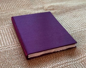 May include: A rectangular, hardcover journal with a deep purple cover. The journal's pages are visible along the edge, and the book is resting on a textured, beige surface. The cover has a subtle fabric texture.