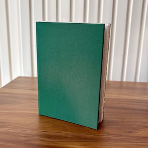 Puede incluir: Un diario de tapa dura verde oscuro est&aacute; de pie sobre una superficie de madera. Las p&aacute;ginas del diario son visibles en el lado derecho, y la cubierta tiene un acabado mate. El fondo presenta una pared de listones blancos.