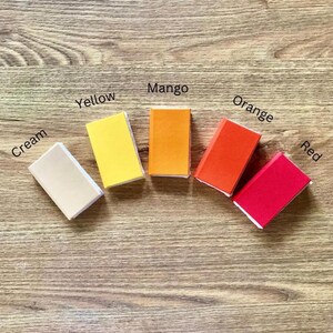 May include: Five small, rectangular objects in cream, yellow, mango, orange, and red are arranged on a wooden surface. Each object has a white border and a colour label: Cream, Yellow, Mango, Orange, and Red.