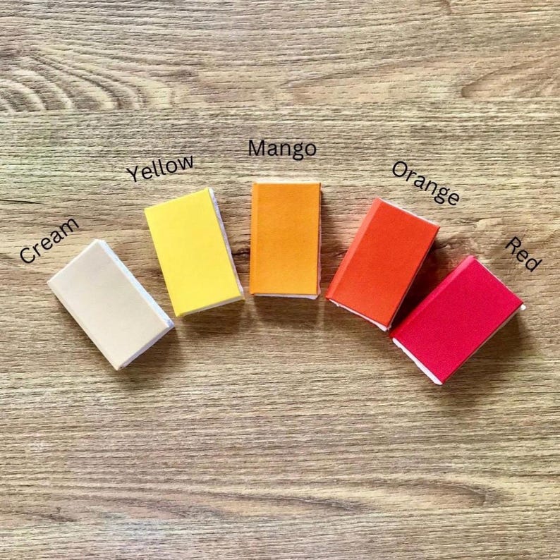 May include: Five small, rectangular objects in cream, yellow, mango, orange, and red are arranged on a wooden surface. Each object has a white border and a colour label: Cream, Yellow, Mango, Orange, and Red.