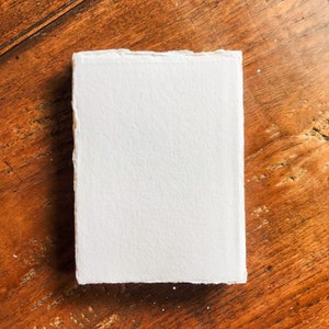 May include: A white, textured, handmade paper sheet with deckled edges. The paper is lying flat on a wooden surface.