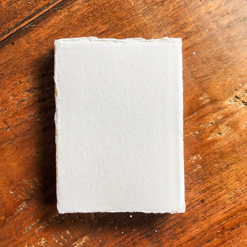 May include: A white, textured, handmade paper sheet with deckled edges. The paper is lying flat on a wooden surface.