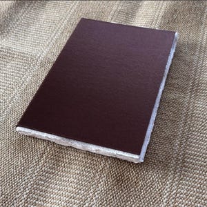 Op de afbeelding: Een rechthoekig, donkerbruin notitieboek met een getextureerde kaft. De pagina's hebben een gescheurde rand, waardoor het een handgemaakte uitstraling heeft. Het notitieboek ligt op een getextureerd, bruin oppervlak.