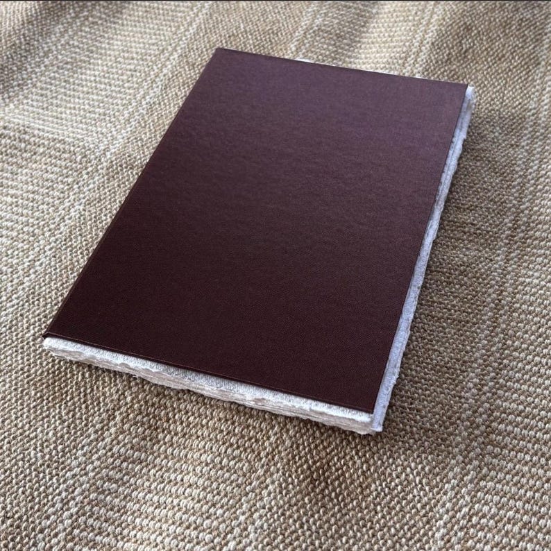 Op de afbeelding: Een rechthoekig, donkerbruin notitieboek met een getextureerde kaft. De pagina's hebben een gescheurde rand, waardoor het een handgemaakte uitstraling heeft. Het notitieboek ligt op een getextureerd, bruin oppervlak.