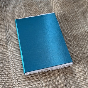 May include: A teal blue notebook with a textured cover. The notebook is closed and lying on a beige fabric surface.