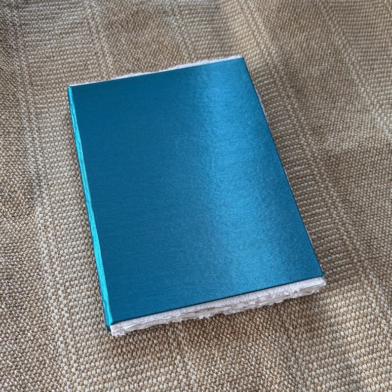 May include: A teal blue notebook with a textured cover. The notebook is closed and lying on a beige fabric surface.
