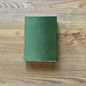 Op de afbeelding: Een klein, rechthoekig hardcover boek met een donkergroene kaft. Het boek is gesloten en ligt op een houten oppervlak. De kaft heeft een licht getextureerde uitstraling.