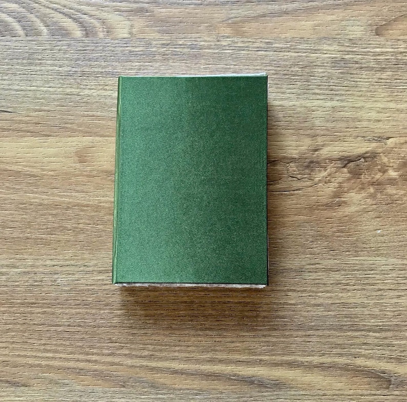 Op de afbeelding: Een klein, rechthoekig hardcover boek met een donkergroene kaft. Het boek is gesloten en ligt op een houten oppervlak. De kaft heeft een licht getextureerde uitstraling.