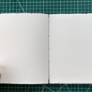 Puede incluir: Un cuaderno de bocetos abierto con p&aacute;ginas texturizadas de color blanco roto. La encuadernaci&oacute;n es visible, con peque&ntilde;as grapas plateadas que aseguran las p&aacute;ginas. El libro est&aacute; abierto sobre una esterilla de corte verde.