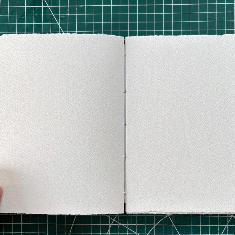Puede incluir: Un cuaderno de bocetos abierto con p&aacute;ginas texturizadas de color blanco roto. La encuadernaci&oacute;n es visible, con peque&ntilde;as grapas plateadas que aseguran las p&aacute;ginas. El libro est&aacute; abierto sobre una esterilla de corte verde.