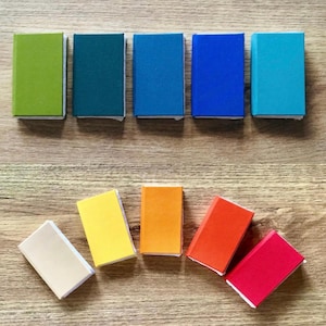 May include: A selection of brightly coloured matchbooks, including shades of green, blue, yellow, orange, and red. The matchbooks are arranged on a wooden surface, displaying their vibrant colours and rectangular shape.