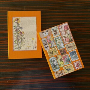 May include: Two orange-colored notebooks. One features a botanical illustration framed within, the other is covered in vintage postage stamp designs, including butterflies and flowers. The notebooks are on a dark wood surface.