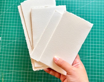 May include: A stack of off-white watercolor paper with a textured surface. The paper has a deckled edge and is held in a hand against a green cutting mat. The paper is suitable for painting and drawing.