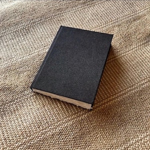 Op de afbeelding: Een gesloten, zwart hardcover boek met een getextureerde kaft. De pagina's zijn zichtbaar langs de rand en het boek ligt op een getextureerd, bruin oppervlak. Het boek lijkt nieuw te zijn.