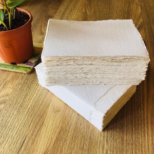 May include: A stack of white handmade paper sheets with a rough, textured surface. The paper is arranged in a rectangular shape and is sitting on a wooden surface.