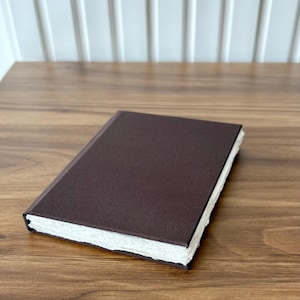 Op de afbeelding: Een gesloten, donkerbruin notitieboek met een getextureerde kaft en gescheurde randen. Het rechthoekige notitieboek lijkt van handgemaakt papier te zijn. De pagina's zijn wit en de kaft is effen donkerbruin. Het notitieboek ligt op een houten oppervlak.