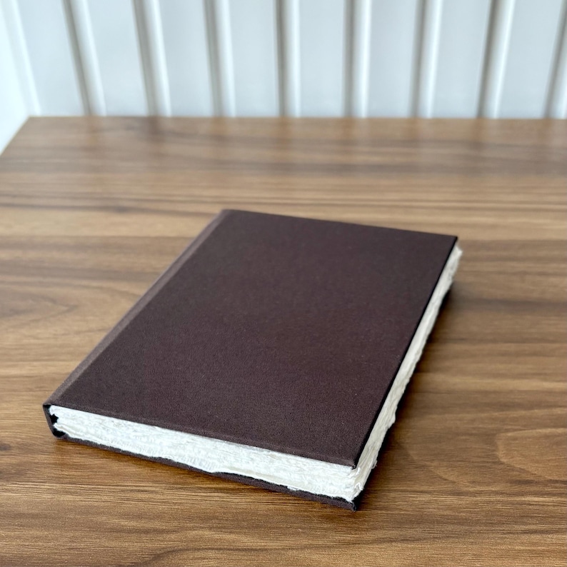 Op de afbeelding: Een gesloten, donkerbruin notitieboek met een getextureerde kaft en gescheurde randen. Het rechthoekige notitieboek lijkt van handgemaakt papier te zijn. De pagina's zijn wit en de kaft is effen donkerbruin. Het notitieboek ligt op een houten oppervlak.