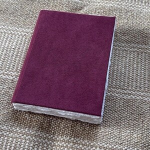 May include: A rectangular notebook with a solid deep burgundy suede-like cover. The edges of the pages are visible along the side, featuring a thick, textured, and deckled white paper finish that contrasts with the smooth, dark-colored cover.