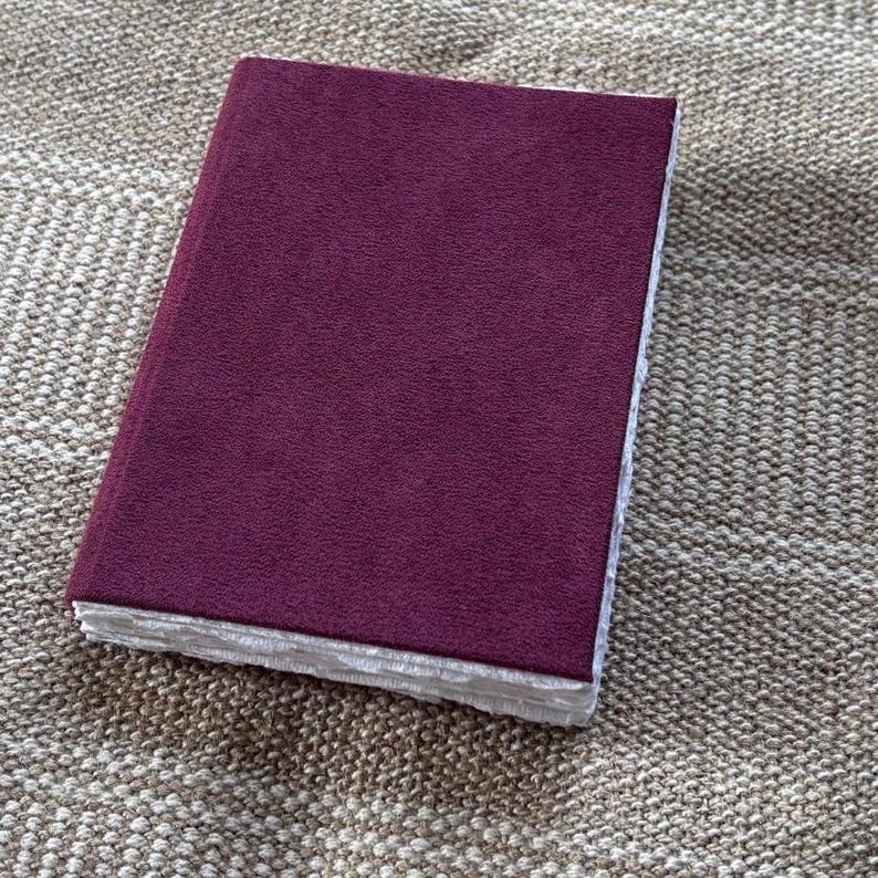 May include: A rectangular notebook with a solid deep burgundy suede-like cover. The edges of the pages are visible along the side, featuring a thick, textured, and deckled white paper finish that contrasts with the smooth, dark-colored cover.