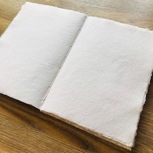 Puede incluir: Un diario abierto y en blanco con p&aacute;ginas texturizadas de color blanquecino. Las p&aacute;ginas se mantienen unidas por una encuadernaci&oacute;n cosida. El diario est&aacute; sobre una superficie de madera, proporcionando un fondo natural.