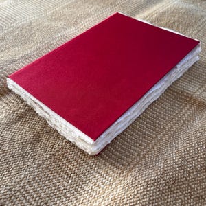 Op de afbeelding: Een rood hardback notitieboek met witte pagina's. Het notitieboek is gesloten en ligt op een getextureerd beige oppervlak.