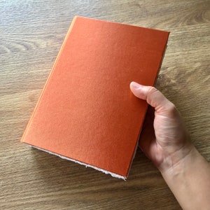 May include: A close-up of an orange notebook with a textured cover. The notebook is being held by a hand.