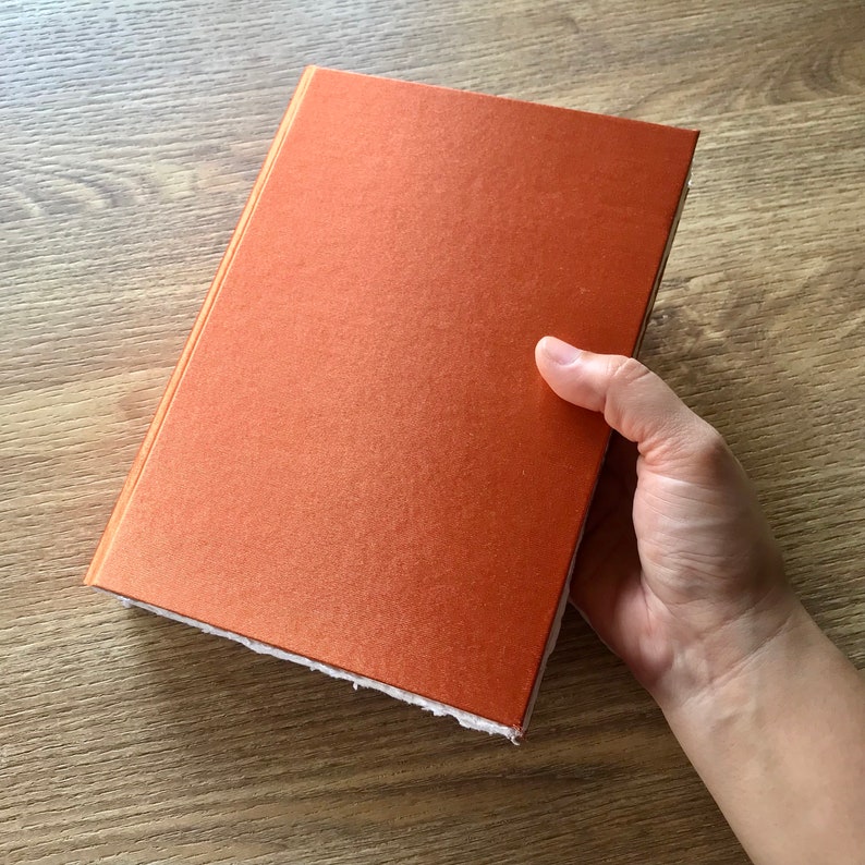 May include: A close-up of an orange notebook with a textured cover. The notebook is being held by a hand.