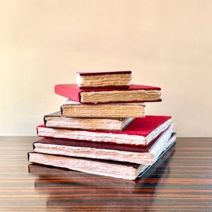 May include: A pile of six handmade journals with red covers and aged, cream-coloured pages. The journals are stacked, with the smallest on top. The edges of the pages are rough-cut, giving them a vintage appearance. The journals are stacked on a wooden surface.