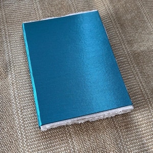 Op de afbeelding: Een turquoise notitieboek met een getextureerde kaft en witte, getextureerde randen. Het rechthoekige notitieboek ligt op een beige, geweven oppervlak. De kaft heeft een lichte glans.